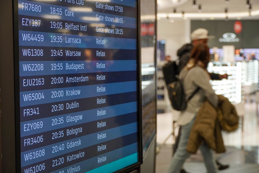Departures boards show flight times. Passengers walk by with rucksacks in background